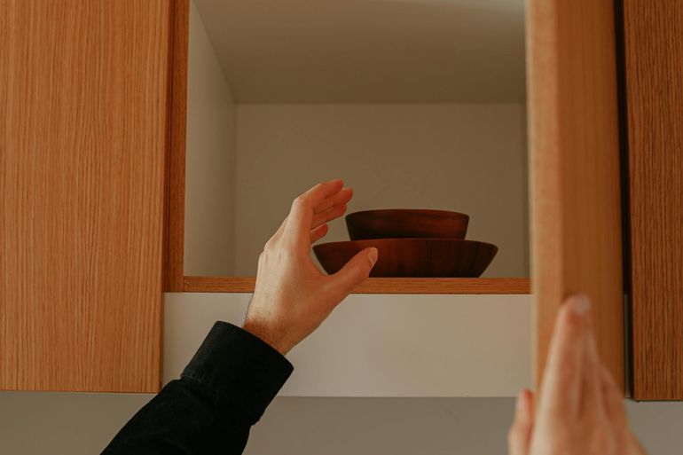 Рука открывает деревянный кухонный шкаф с посудой внутри